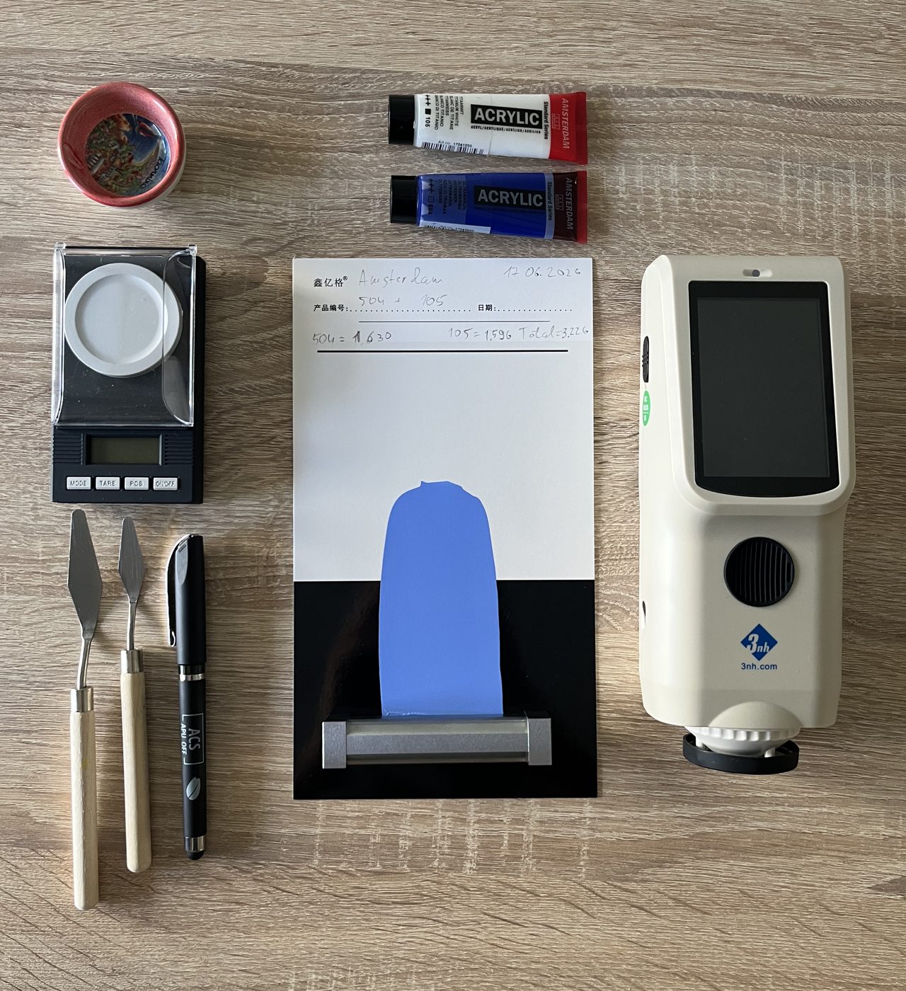 A bench laid out with a precision scale, paint tubes, mixing dish, palette knives, an opacity card and a spectrophotometer.